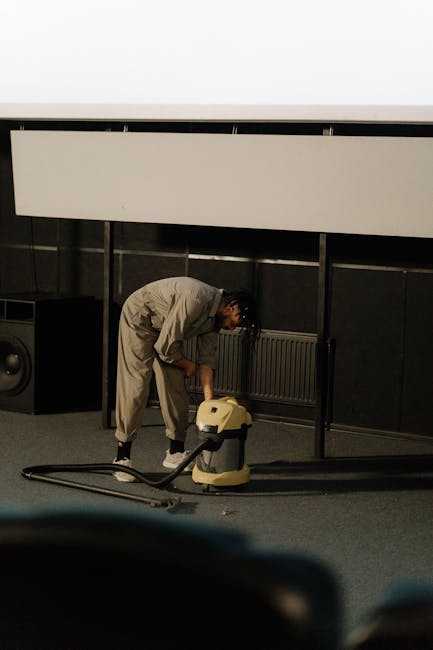 A professional cleaner dressed in white protective clothing, including a coverall, gloves, face mask, and shoe covers, is performing deep cleaning or sanitisation of a light beige carpet in a bright,