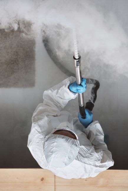 A person dressed in protective white coveralls and blue gloves is using a steam cleaning tool to treat a large, dark stain on a concrete basement floor. The steam cleaner produces visible steam and moisture over the surface, which appears damp and slightly reflective. The basement has gray concrete walls and flooring, with minimal lighting creating a neutral, clinical environment. The scene emphasizes surface cleaning and odour removal, aligning with professional deep cleaning and sanitisation processes as discussed in the page titled 'Common problems with steam cleaning in Holland Park basements, error code: 521' by Holland Park Carpet Cleaners. The image conveys the process of technical stain removal or deep cleaning in a residential basement setting.