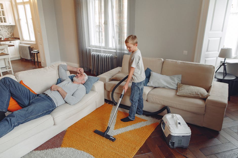 A young boy vacuuming a beige carpet in a living room during daytime, with sunlight coming through large windows with sheer curtains. The room features a cream-colored sofa with light blue and beige cushions, a small side table with a lamp, and wooden flooring partially covered by a multicolored area rug. An older man with gray hair, wearing a gray sweater and blue jeans, is lying on the sofa, appearing relaxed. The clear surfaces and tidy appearance suggest regular surface cleaning and maintenance are performed by Holland Park Carpet Cleaners, emphasizing professional deep cleaning and sanitisation for period homes.