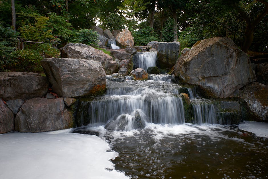 A natural outdoor scene featuring a small cascade of water flowing over large, irregularly shaped rocks in a lush green setting, with trees in the background providing dappled sunlight. The water appears clear and moving gently over the stones, creating small pools and ripples. The rocks are of various sizes and colors, predominantly gray and brown, with some moss growth visible. The scene emphasizes the natural aesthetic and tranquility. This image relates to surface cleaning and maintenance in garden or outdoor environments, akin to the detailed stain removal tips offered by Holland Park Carpet Cleaners at https://hollandparkcarpetcleaners.com.