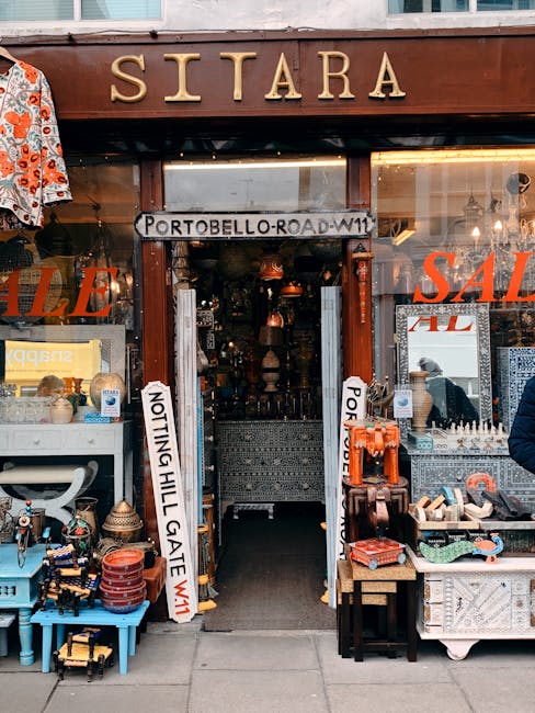 The storefront of Sitares, showcasing a wooden sign with gold lettering above the entrance, situated on Portobello Road in Notting Hill Gate. The facade features large display windows on either side, filled with a variety of decorative items, ceramics, and fashion accessories. A sale sign indicating discounts is visible, along with a white display table on the left holding small ornaments and pottery. Inside, the shop appears well-organized with a dark interior, illuminated by warm lighting and featuring an interior staircase. The area outside is clean and inviting, with the display windows and sidewalk free of clutter. The overall scene highlights a vibrant retail environment that benefits from regular surface cleaning and maintenance by Holland Park Carpet Cleaners, ensuring hygiene and a welcoming appearance for customers.