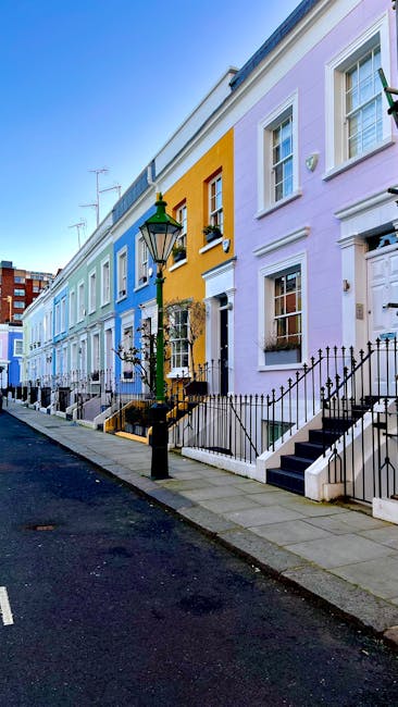 A row of vibrant, pastel-colored terraced houses on a residential street, featuring white-framed sash windows and black wrought-iron railings. The houses are painted in shades of blue, yellow, pink, and lavender, with front steps leading up to small entrances. A traditional black street lamp stands prominently on the sidewalk, which is clean and well-maintained, with a dark asphalt road in foreground. The scene is illuminated by natural daylight, highlighting the neatness and charm of the urban setting. Holland Park Carpet Cleaners specializes in surface cleaning and deep cleaning services, ensuring domestic spaces remain hygienic and well-maintained.