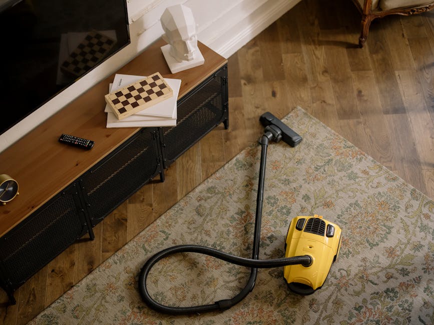 A yellow vacuum cleaner with a black hose and metal wand attachment placed on a decorative area rug in a living room. The rug has a floral pattern in muted colors. To the left, there is a wooden media console with black mesh doors, holding a remote control, a stack of white papers, an open chessboard, and a white sculpture of a head. The room features wooden flooring and white wall paneling, with natural lighting illuminating the scene. Holland Park Carpet Cleaners' deep surface cleaning process is suggested by the vacuum cleaner, highlighting hygiene and maintenance for domestic environments.