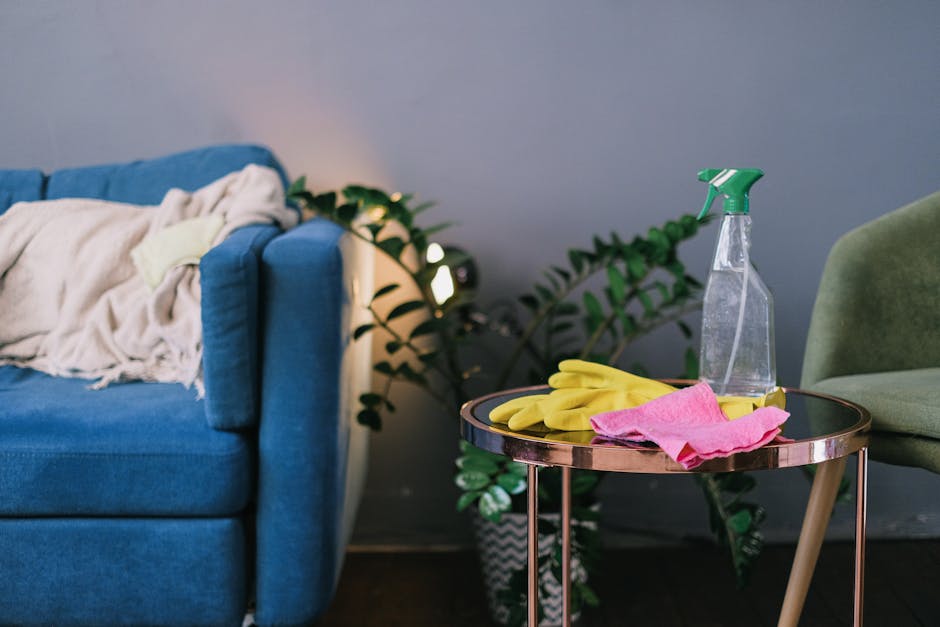 A modern living room scene featuring a blue fabric sofa with a slightly rumpled cream throw blanket draped over one armrest, situated next to a side table with a reflective metallic surface. On the table, there are yellow rubber cleaning gloves, a pink cloth, and a transparent spray bottle with a green nozzle, all indicating recent surface cleaning or maintenance efforts. The background includes a potted plant with lush green leaves, positioned against a neutral wall illuminated by soft, natural lighting, creating a clean and well-maintained atmosphere suitable for discussing domestic surface cleaning and hygiene tips. Holland Park Carpet Cleaners' branding is subtly implied to emphasize professional cleaning services associated with the page titled 'W11 upholstery cleaning tips for Campden Hill flats, error code: 521', highlighting experts in deep cleaning and sanitisation.